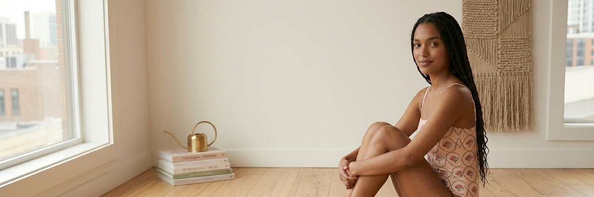 Model in a floral-print one-piece sitting on the floor of a bright room beside a brass watering can